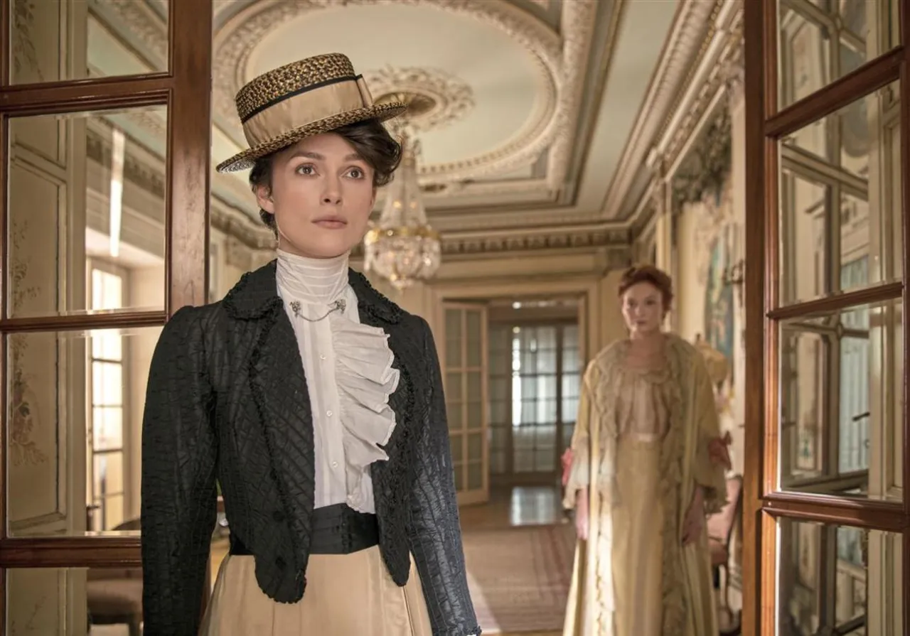 Dos mujeres con vestuario históricamente correcto en un pasillo lujoso. La de adelante, con sombrero, luce un traje negro y falda beige.
