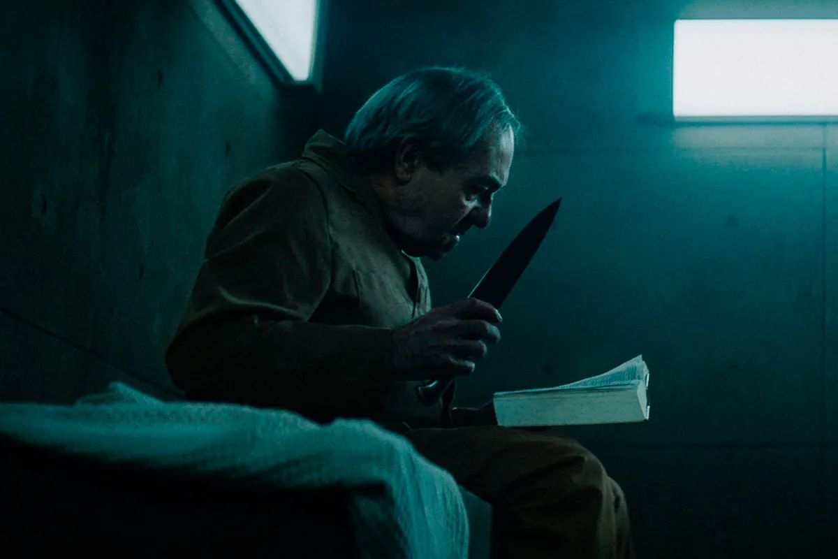 Un hombre con un cuchillo y un libro, en una escena que evoca la explicación detallada del final de la película El Hoyo.