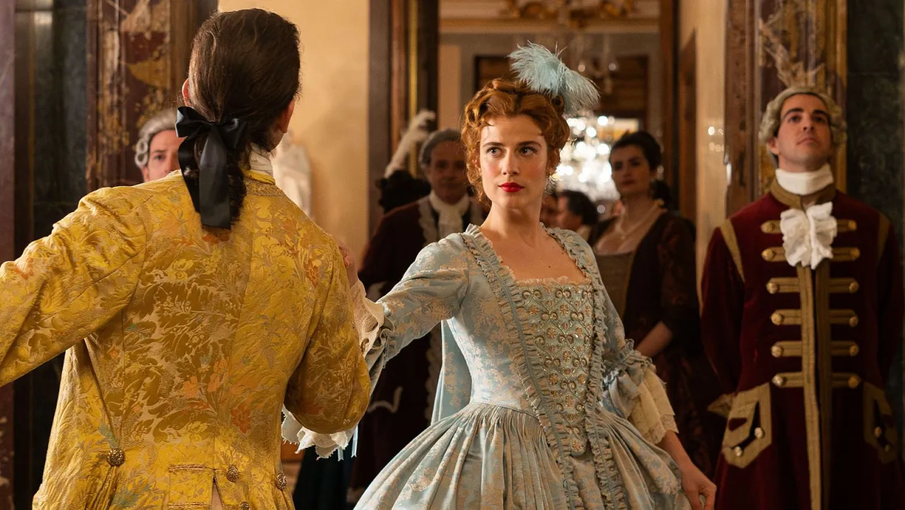 Una pareja baila en un salón, luciendo vestuarios de películas de época con vestuario históricamente correcto. Ella, con un vestido azul claro, él, con una chaqueta dorada.
