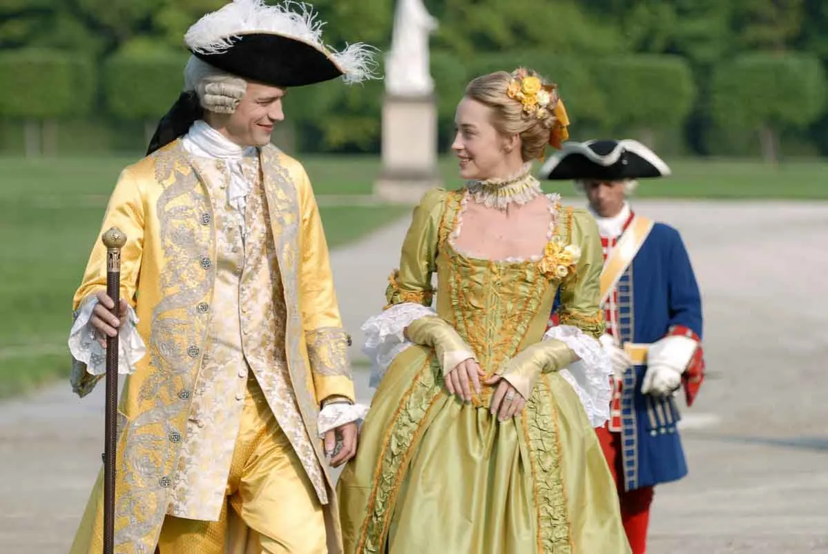 Pareja con vestuario históricamente correcto para películas de época, paseando por un jardín. Él lleva un traje amarillo con bordados, ella un vestido verde con volantes.